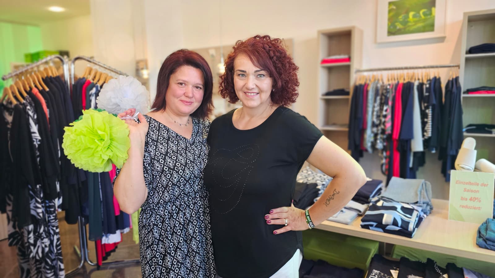 Anett Müller und Lena Miller stehen in der Boutique Schwungvoll mit einem grünen Ponpon und werben für Ihre Modenschau